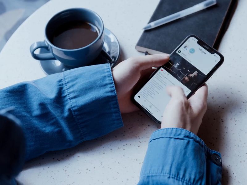 osoba u kafiću lista instagram, pored nje šoljica sa kafom i rokovnik sa hemijskom